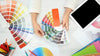 A designer holding paint color chips fanned out on a table with a tablet and other design tools.